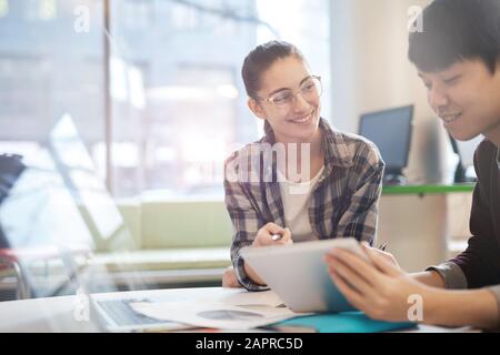 Young woman in eyeglasses sitting at the table pointing at tablet pc and smiling she talking to her colleague at office Stock Photo