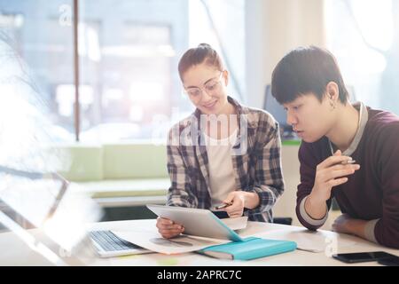 Young woman in eyeglasses pointing at digital tablet and discussing online work with her colleague while they sitting at the table Stock Photo