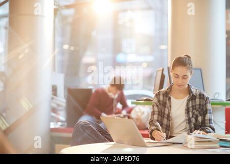 Young woman sitting at the table in front of laptop and making notes in notebook she working in the library Stock Photo