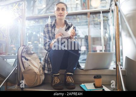 Young woman sitting on staircase with schoolbag and laptop and making notes in her notepad she preparing for lessons Stock Photo
