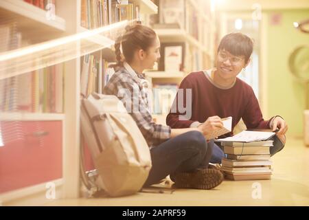 Two friends talking and smiling to each other while sitting on the floor with heap of books and studying them Stock Photo