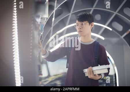 Asian young man in eyeglasses standing near the bookshelf and choosing books for reading in the library Stock Photo