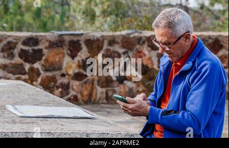 Older man looking and touching the smartphone screen with the newspaper on the table while sitting in the park Stock Photo