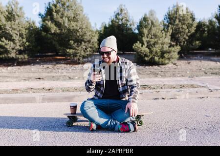 Modern young man with a wool hat and sunglasses laughing while sitting on the longboard looking at his mobile phone Stock Photo