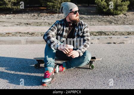 Young modern man with a wool hat and sunglasses sitting on the longboard having a coffee Stock Photo