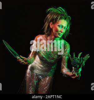 Studio image of young woman wrapped in plastic film and holding a knife a bunch of green vegetables. Stock Photo