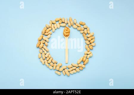 Creamy peanut paste in wooden spoon. Peanuts in the peel scattered on blue background. Minimalistic food flat lay on color blue background. Stock Photo