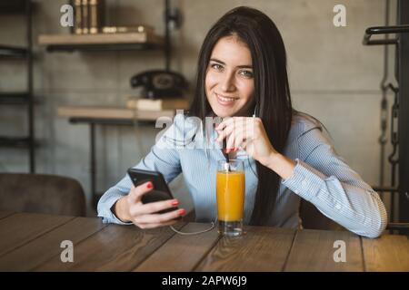 Positive young woman in casual clothes drinking fresh beverage using phone sitting at table in cafe Stock Photo