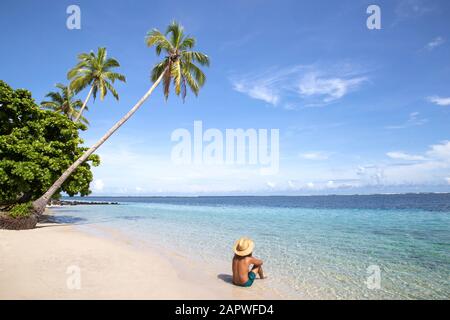 Man with golden hat and green swimsuit at beach with leaning palm tree Stock Photo