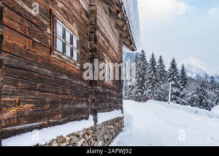 Wooden wall of mountain house, against the backdrop of snowy mountains and trees. Winter alpine landscape in Austria. Beautiful natural background. Stock Photo