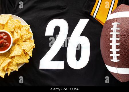 An American football with organic nacho chips and mild salsa on a white black football jersey with the 26 number on horizontal view Stock Photo