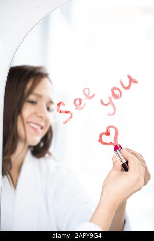 Hand of happy female writing see you and drawing heart with lipstick on mirror Stock Photo