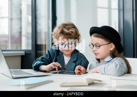 Cute elementary school friends in casualwear and eyeglasses using digital tablet Stock Photo