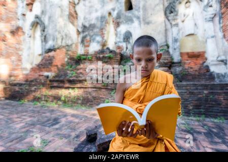 Novice monk reading outdoors, sitting outside monastery, Thailand Stock Photo