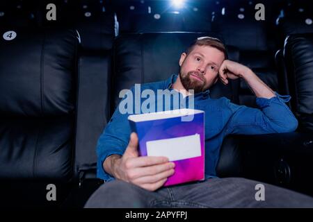 Young bored man with box of popcorn sitting on black leather armchair Stock Photo