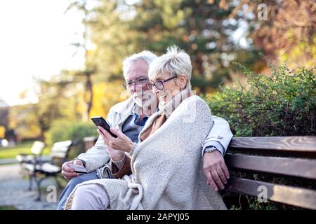 Senior couple shopping on line with mobile phone and credit card while sitting on the bench in park Stock Photo
