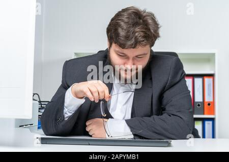 a stressed employee sits at the desk and holds his glasses in his hand Stock Photo