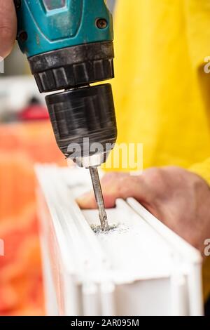 Male hands with an electric screwdriver and drill make a hole in the PVC window frame. Stock Photo