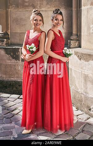 Two beautiful bridesmaids girls blonde and brunette ladies wearing elegant full length red chiffon bridesmaid dress with lace and holding flower Stock Photo