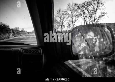 Female photographer taking a photo of herself in the right rearview mirror of a moving car driving in a conventional road. Black and white. B&W.Selfie Stock Photo