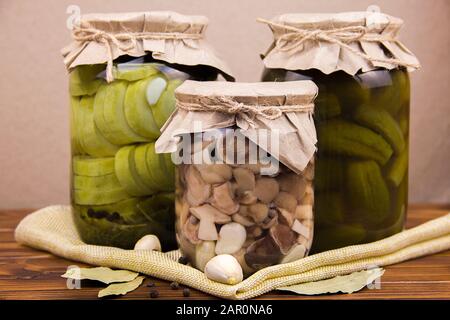 fermented pickled canned zucchini and cucumber mushrooms in jars garlic bay leaf black pepper peas on a piece of burlap on a brown wooden background Stock Photo