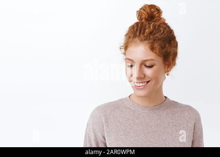Lovely, attractive and tender redhead curly woman in stylish blouse, lookign down, blushing from compliment, standing happy and modest, smiling Stock Photo