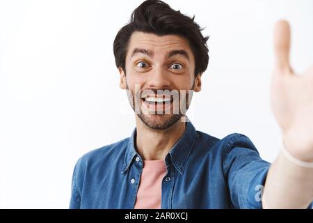 Cheerful, friendly young man with beard, trendy haircut taking selfie on smartphone, hold camera with raised hand, smiling, making joyful expression Stock Photo