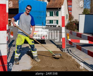 Laughing construction worker with broom standing on sand in front of vehicle and trailer looking into camera. Stock Photo
