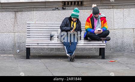London, UK - January 1, 2020: Two men sit on a bench on the side of the street and look at their smartphones Stock Photo