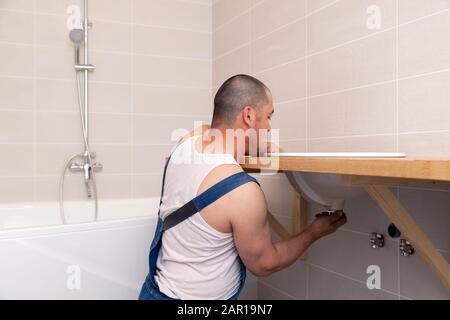 Closeup male plumber worker in blue denim uniform, overalls, fixing sink in bathroom with tile wall. Professional plumbing repair service, installatio Stock Photo