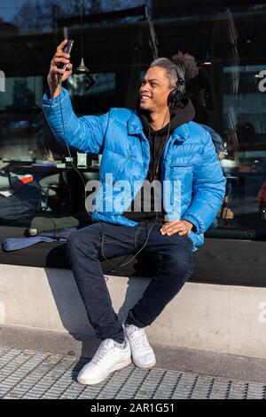 young afroamerican is making a video call with smartphone and headphones while sitting in front of a coffee shop, lifestyle concept using new technolo Stock Photo