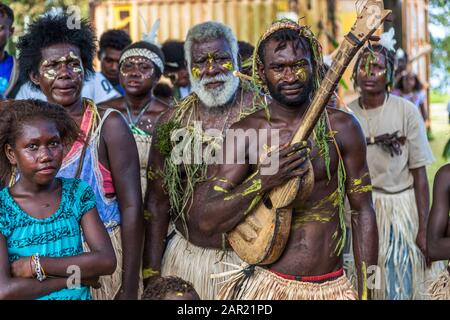 A man with yellow body paint plays a hand-carved stringed instrument at a Sing-Sing village festival in Bougainville, Papua New Guinea, beside him an older, white-bearded man wears a necklace of green leaves. Stock Photo