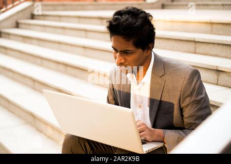 Young handsome Indian man is sitting with a laptop, smiling and typing Stock Photo