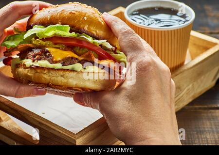 Woman's hands holding a hamburger over wooden tray and paper cup of drink