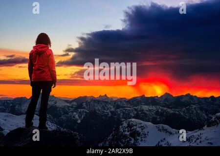 Adventurous Girl watching the Beautiful Dramatic Sunset on top of the Mountains. Composite Image with Landscape taken in British Columbia, Canada. Con Stock Photo