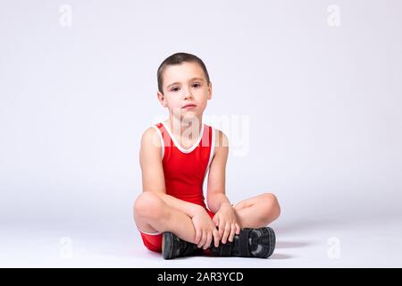 A boy athlete in sportswear and wrestling outfits sits on the floor and looks sadly at the camera on a white isolated background. Stock Photo