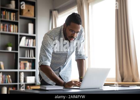 Focused african businessman standing at home office desk using laptop Stock Photo