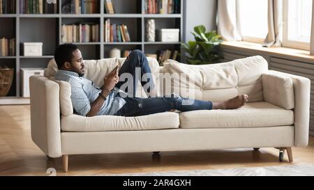 Serious relaxed millennial african man lying on couch using smartphone Stock Photo