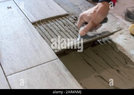 Man installing rectangular shaped floor tiles in kitchen. Applying adhesive before installation and verifying afterwards Stock Photo