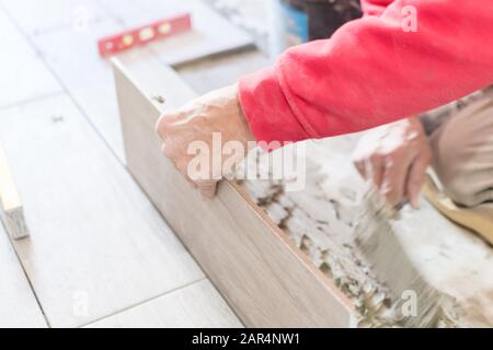Man installing rectangular shaped floor tiles in kitchen. Applying adhesive before installation and verifying afterwards Stock Photo