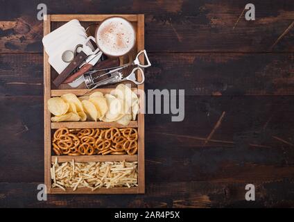 Glass of craft lager beer in vintage box of snacks openers and beer mats on wood background. Pretzel and crisps and salty potato. Stock Photo