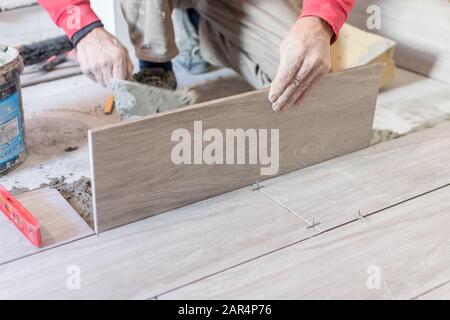 Man installing rectangular shaped floor tiles in kitchen. Applying adhesive before installation and verifying afterwards Stock Photo