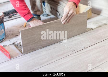 Man installing rectangular shaped floor tiles in kitchen. Applying adhesive before installation and verifying afterwards Stock Photo