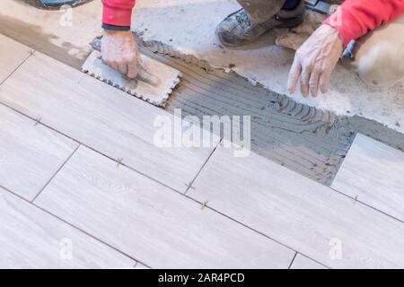 Man installing rectangular shaped floor tiles in kitchen. Applying adhesive before installation and verifying afterwards Stock Photo