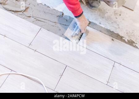 Man installing rectangular shaped floor tiles in kitchen. Applying adhesive before installation and verifying afterwards Stock Photo
