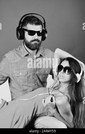 Relax, party and music concept. Music fans with serious faces enjoy music. Man with beard holds girl lying on green and pink background. Couple in love wears headphones and sunglasses. Stock Photo