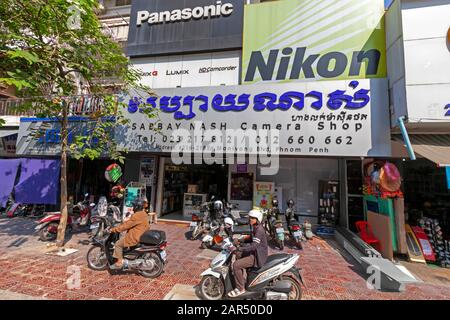 People are riding motorcycles on a sidewalk in front of a retail camera store in Phnom Penh, Cambodia. Stock Photo