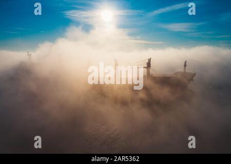 Cargo ship and cranes silhouettes in sea fog, crane vessel working for delivery of delivery containers. Stock Photo