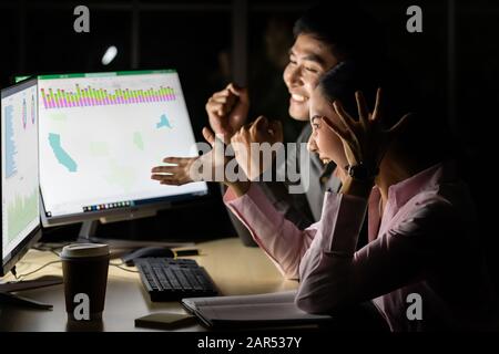 Attractive cheerful glad coworkers gathering celebrating successful work at night at workplace station in dark room with light from desktop computer. Stock Photo
