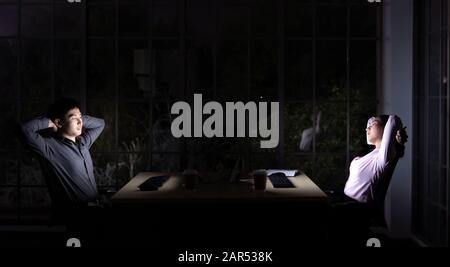 Young adult asian businessman and woman stretching while working late at night in their office with desktop computer and laptop. Using as hard working Stock Photo